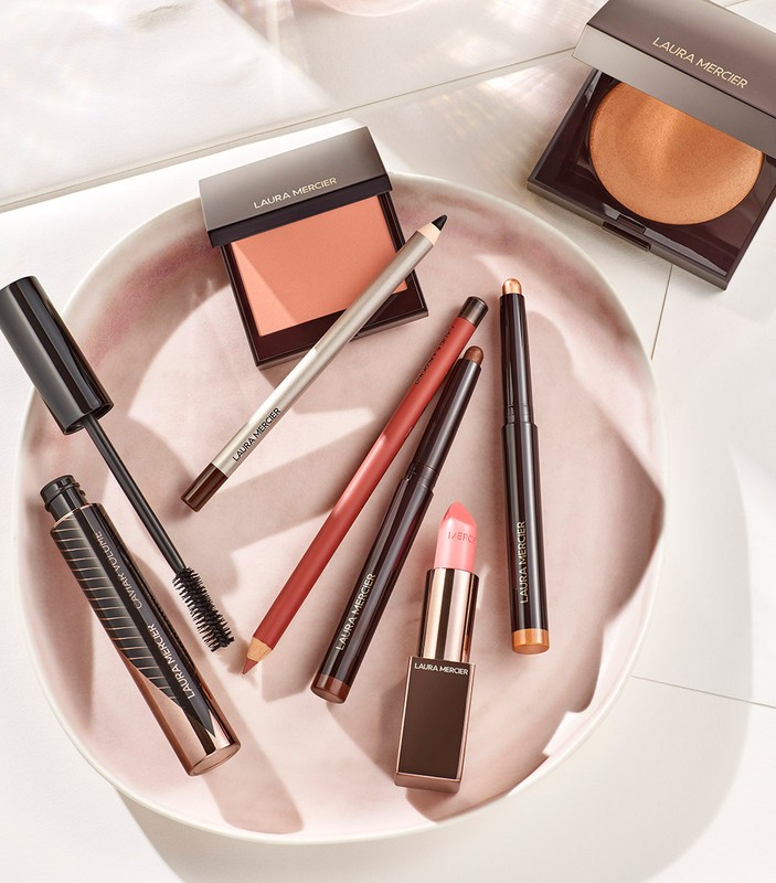Laura Mercier cosmetics arranged on a pink tray, including mascara, blush, eyeliners, and lipstick, with a clean, bright product shot.