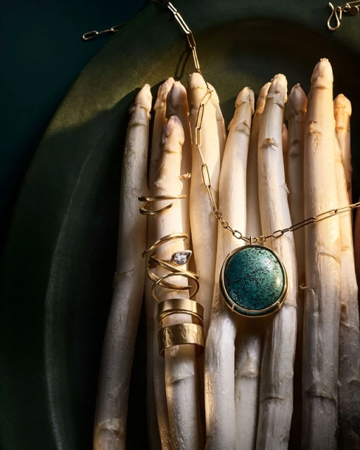 Still life photograph of a gold pendant and rings on a bed of white asparagus against a dark green background with soft studio lighting.