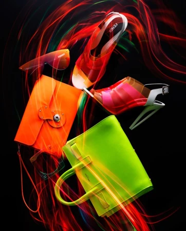 Fashion still life featuring designer handbag, sunglasses, and stilettos against a black background with red and orange light painting.