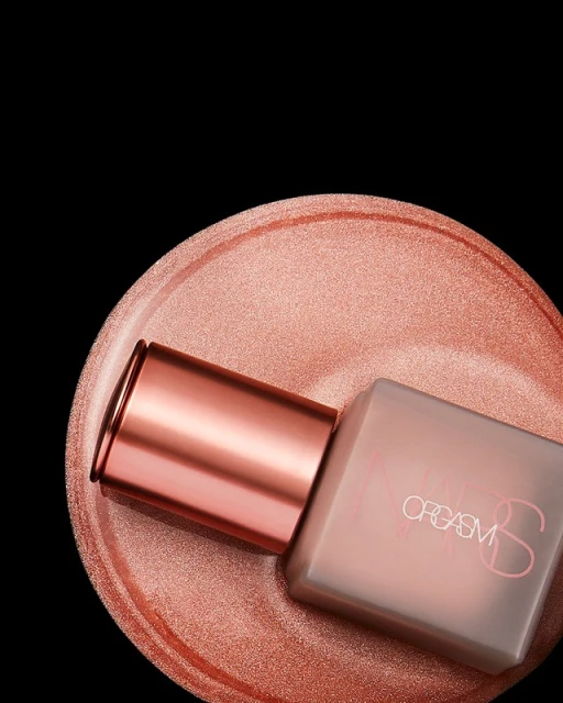 Close-up shot of a NARS Orgasm product bottle sitting on a shimmery peach-colored surface against a stark black backdrop.