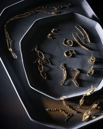 Overhead shot of various gold jewellery pieces including earrings and necklaces arranged on dark octagonal trays.