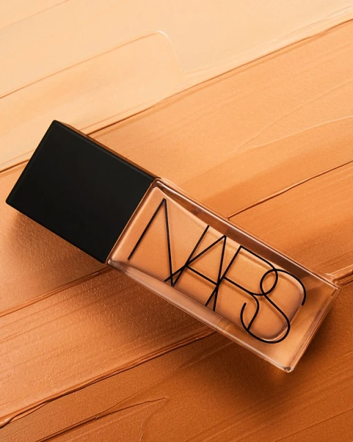 Close-up of NARS foundation bottle against a textured, streaked background of peach and beige foundation swatches, showcasing makeup.