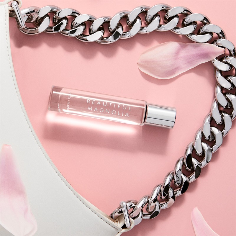 Close-up of a clear, glass perfume bottle of Beautiful Magnolia next to a white purse with chain and magnolia petals on a pink surface.