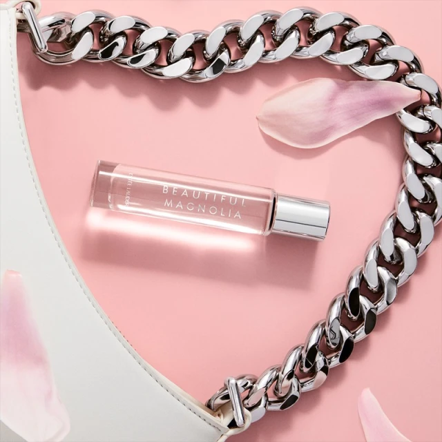 Close-up of a clear, glass perfume bottle of Beautiful Magnolia next to a white purse with chain and magnolia petals on a pink surface.