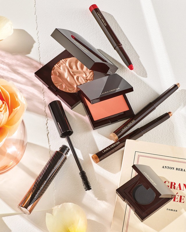 Still life shot of Laura Mercier makeup products including lip crayon, eye shadow, mascara, and powder compacts arranged on a white surface.