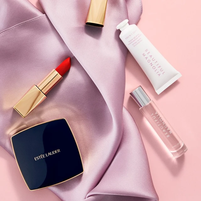 Cosmetic products including lipstick, compact, lotion, and perfume on a pink and purple satin fabric background in a flat lay composition.