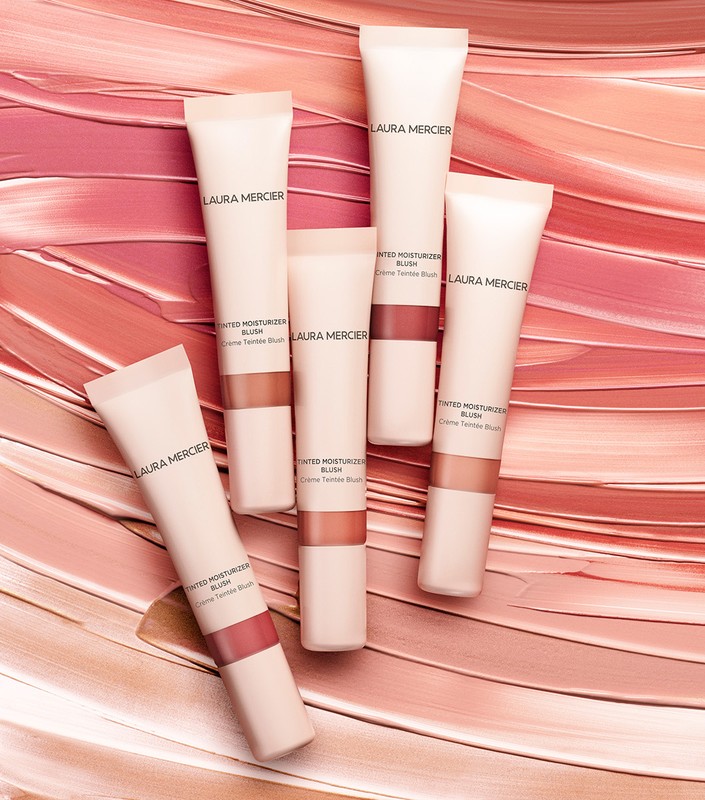 Laura Mercier Tinted Moisturizer Blush tubes are arranged on a background of blush color streaks, showcasing beauty and cosmetics.