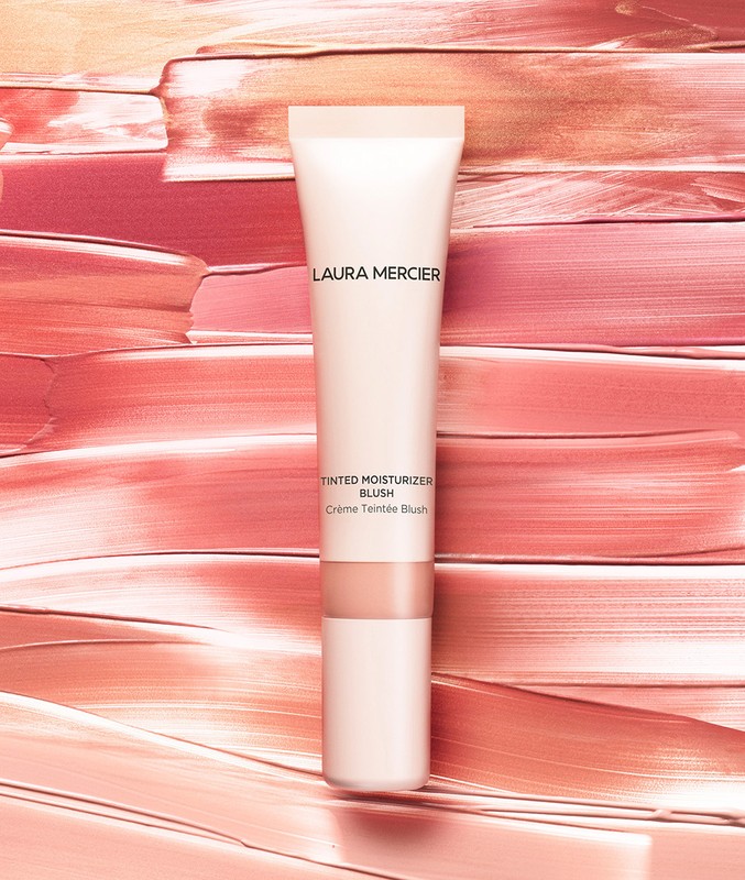 Close-up of Laura Mercier tinted moisturizer blush product in a tube against a textured background of peachy pink strokes.