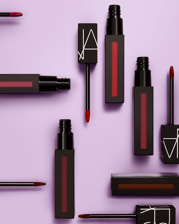 Flat lay of NARS brand lip gloss products in black square containers, arranged against a lavender background in a studio setting.