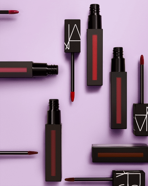 Flat lay of NARS brand lip gloss products in black square containers, arranged against a lavender background in a studio setting.