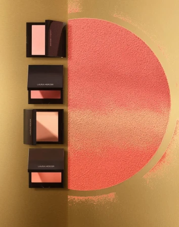 Laura Mercier bronzing powders on a gold background, with semi-circle of loose powder of the same tone, studio lighting.