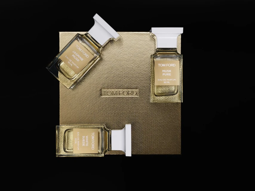 Three Tom Ford perfume bottles on a gold patterned box, set against a stark black background, showcasing a luxurious fragrance presentation.