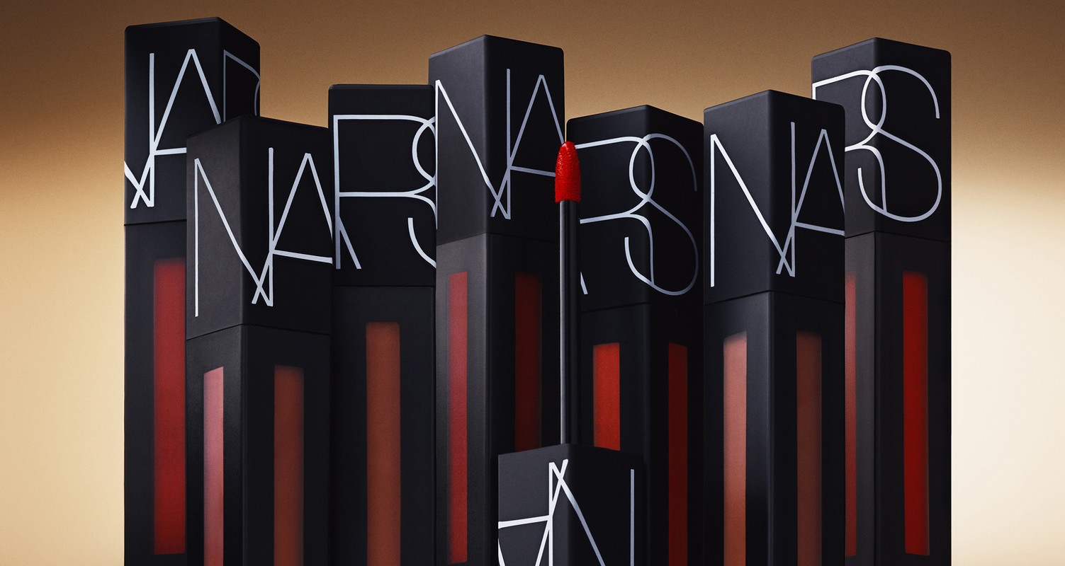 NARS brand lipsticks are stacked in a studio product shot, showcasing their matte black cases and vibrant colors against a tan backdrop.