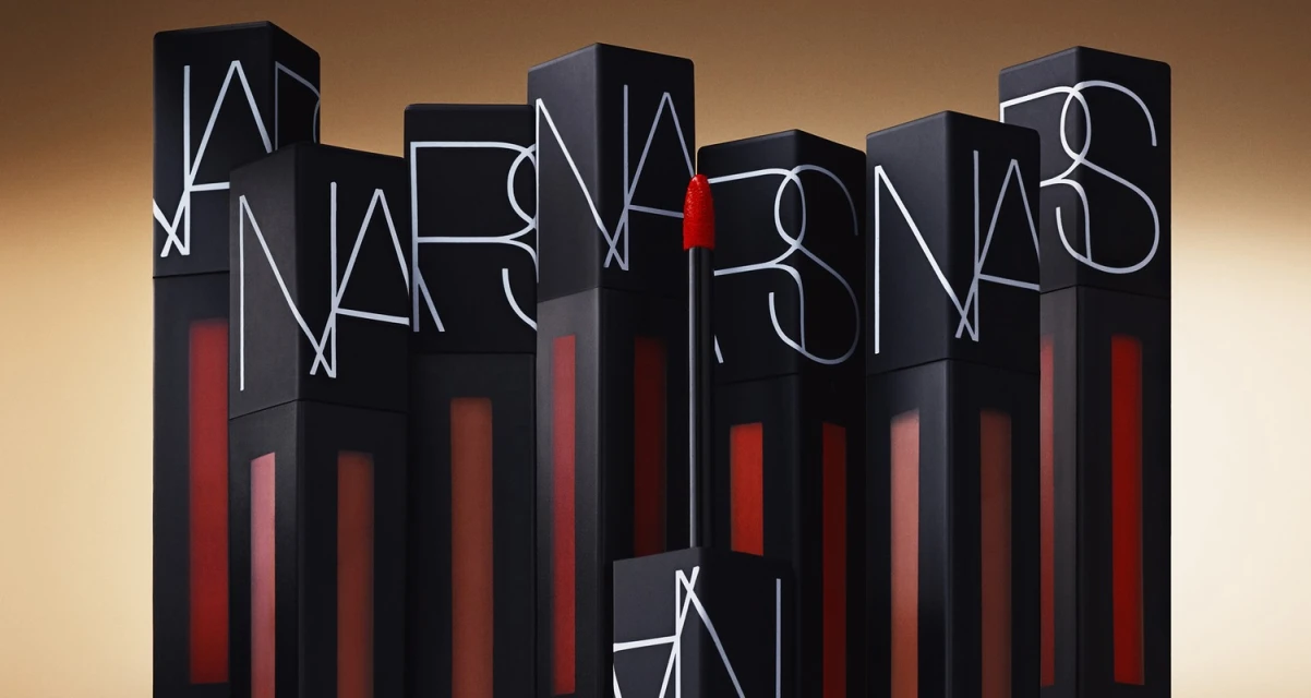 NARS brand lipsticks are stacked in a studio product shot, showcasing their matte black cases and vibrant colors against a tan backdrop.