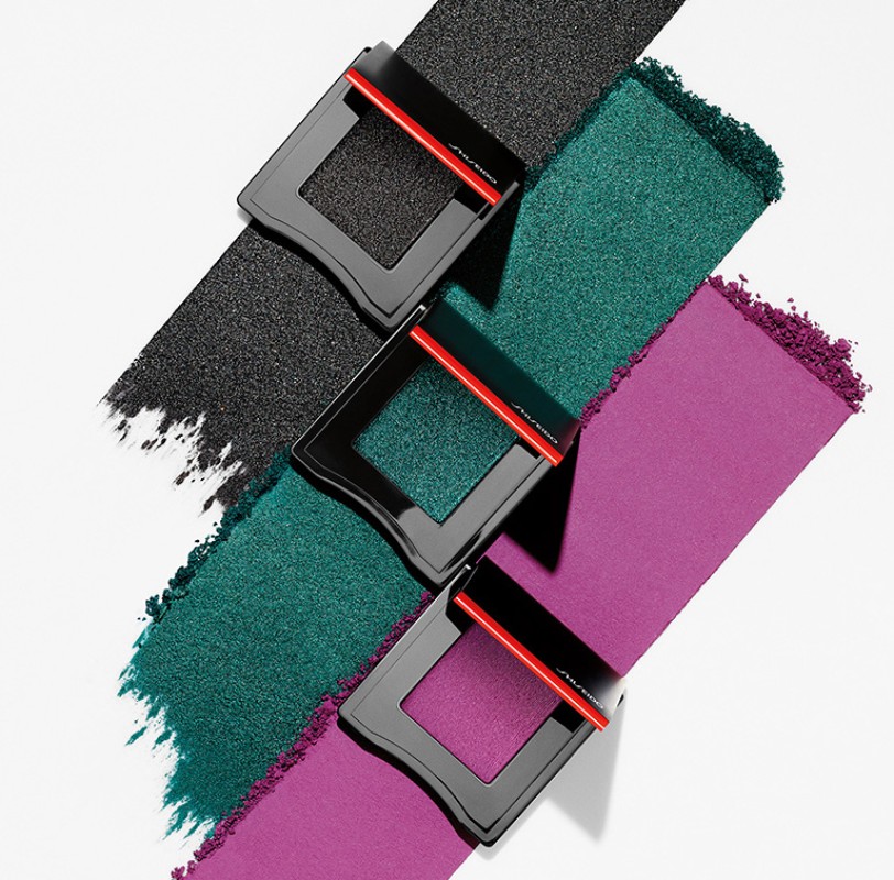 Three square compacts featuring black, teal, and purple powder arranged diagonally on a white surface. Makeup and beauty still life.