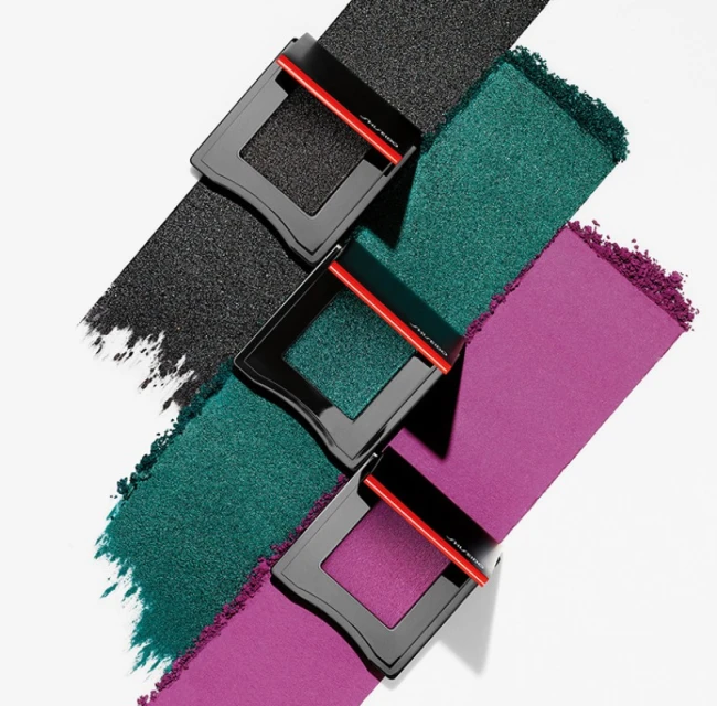 Three square compacts featuring black, teal, and purple powder arranged diagonally on a white surface. Makeup and beauty still life.