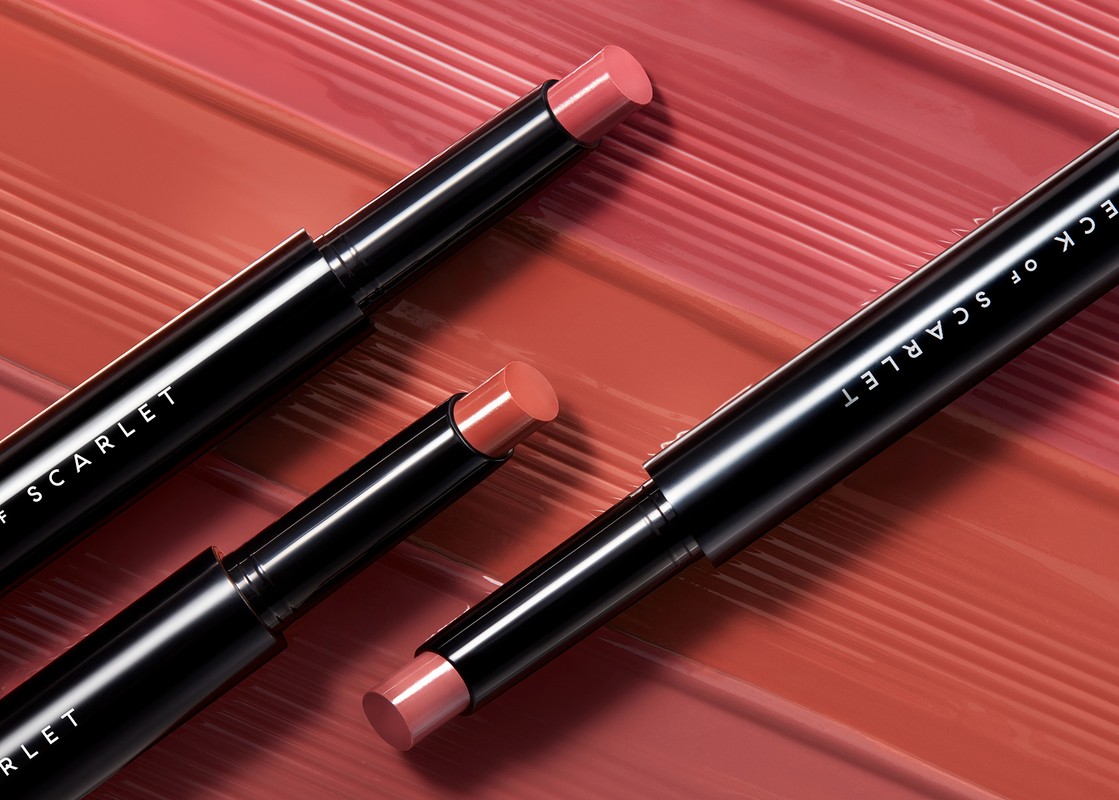 Three Deck of Scarlet lipstick tubes are displayed on a glossy background in coral, showcasing the product's sleek design and color.