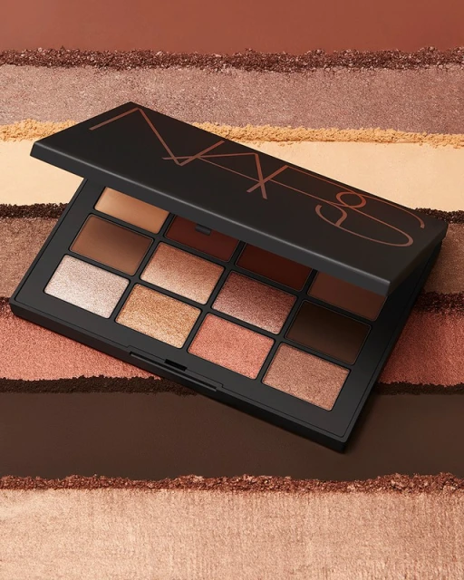 NARS eyeshadow palette with various neutral, earthy tones, set against a background of textured powders. Beauty and makeup product shot.