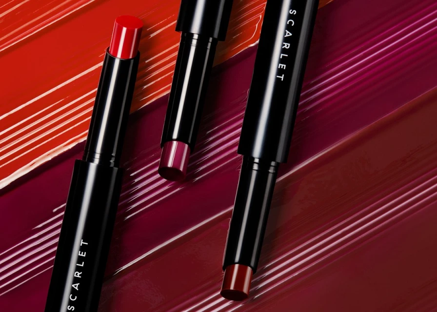 Three Scarlet brand lipsticks in red and burgundy shades are displayed against a matching background of smeared lipstick streaks.