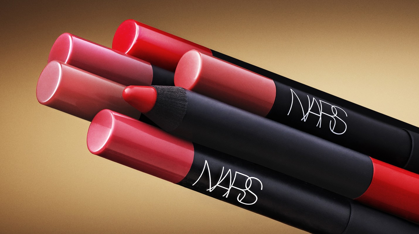 Closeup of NARS lip crayons in red and pink hues arranged diagonally on a tan background. Beauty product photography.
