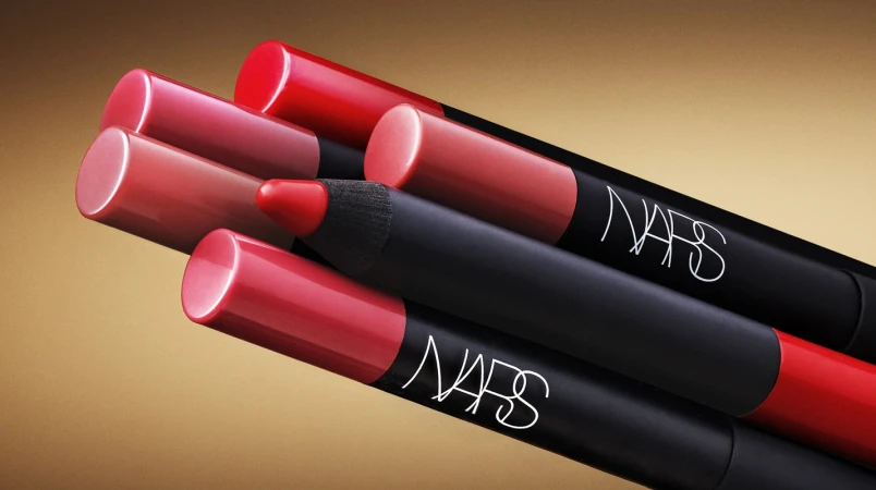 Closeup of NARS lip crayons in red and pink hues arranged diagonally on a tan background. Beauty product photography.