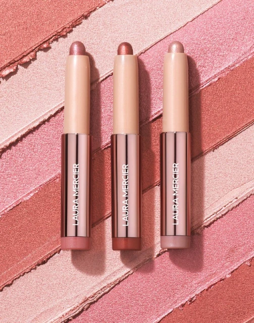Three Laura Mercier caviar eye shadow sticks are displayed against a background of shimmery eyeshadow swatches in pink and peach tones.