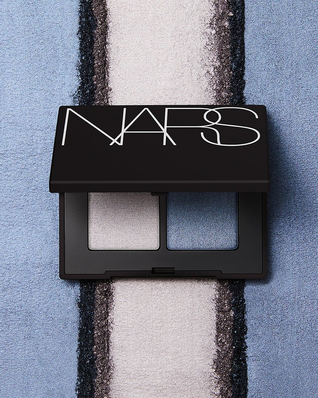 Overhead studio shot of a NARS eye shadow palette featuring silver and blue shimmer shades against a textured blue background.