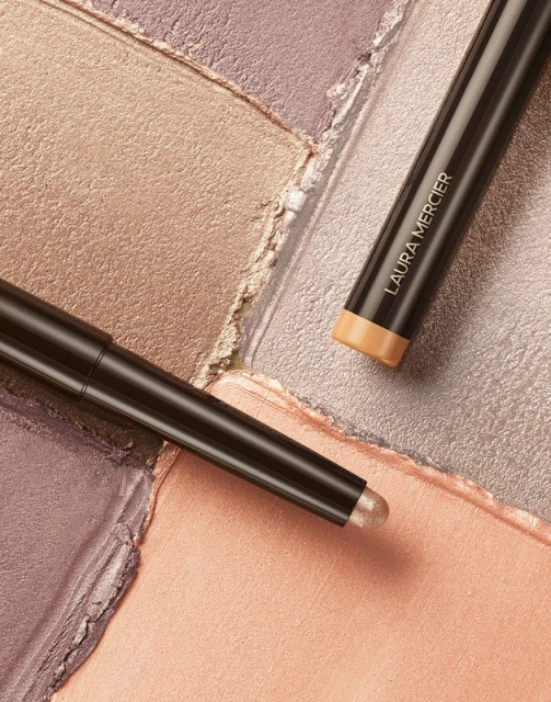 Laura Mercier eye color sticks on shimmering eyeshadow palette in peach, gold, lavender, and silver tones, makeup product shot.
