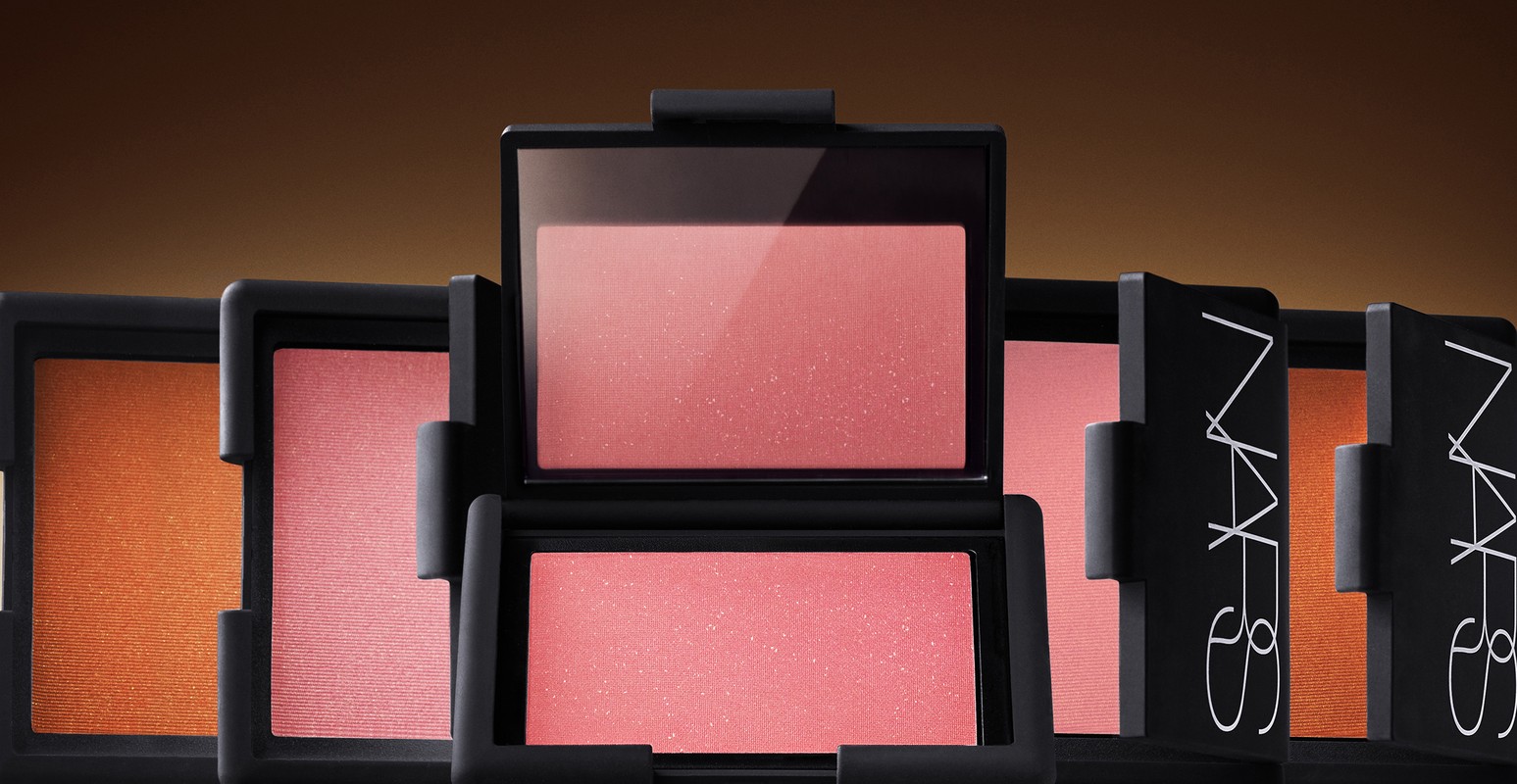 Several blush compacts by NARS are arranged in a row, featuring varying shades of pink and orange, set against a warm brown background.