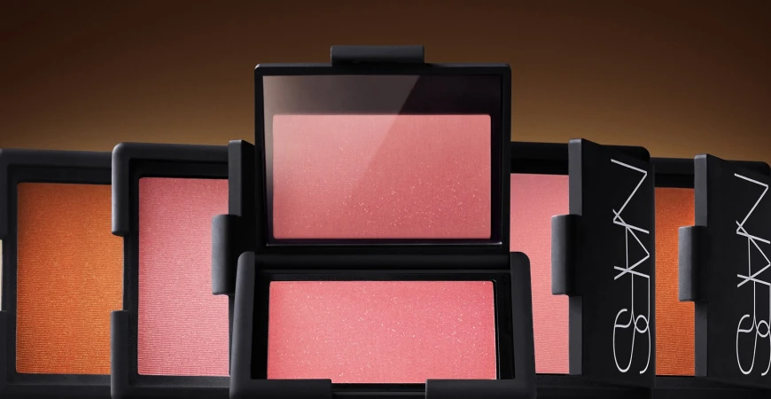 Several blush compacts by NARS are arranged in a row, featuring varying shades of pink and orange, set against a warm brown background.