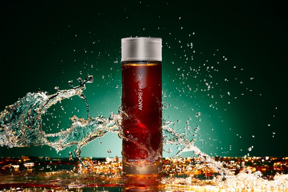 Amore Pacific bottle with splash of water on glass surface against teal studio backdrop in a beauty product shot for cosmetics industry.