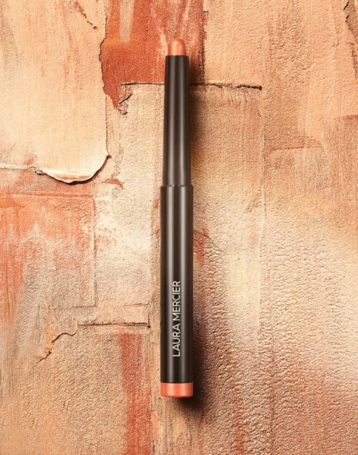 Laura Mercier eyeshadow stick on a textured surface. Product features a warm, shimmering coral shade against a neutral-toned backdrop.