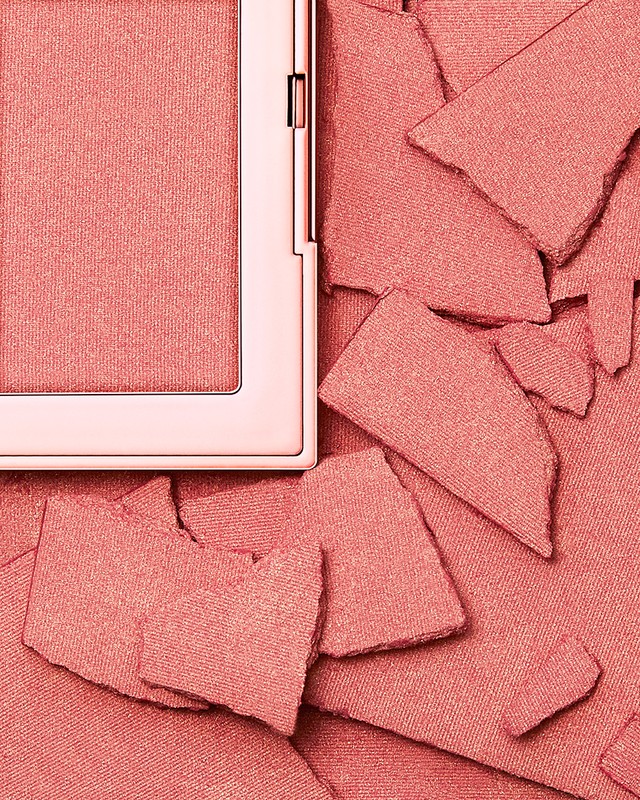 Close-up studio shot of coral-colored blush powder in its compact alongside broken pieces of the same powder for beauty and cosmetic usage.