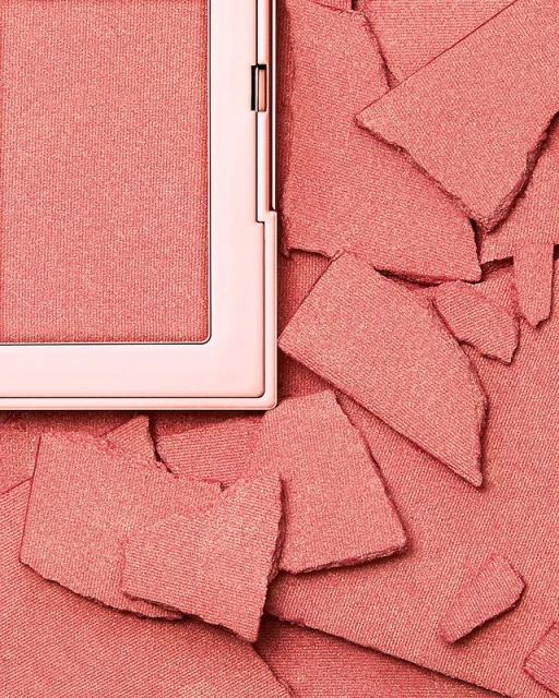 Close-up studio shot of coral-colored blush powder in its compact alongside broken pieces of the same powder for beauty and cosmetic usage.