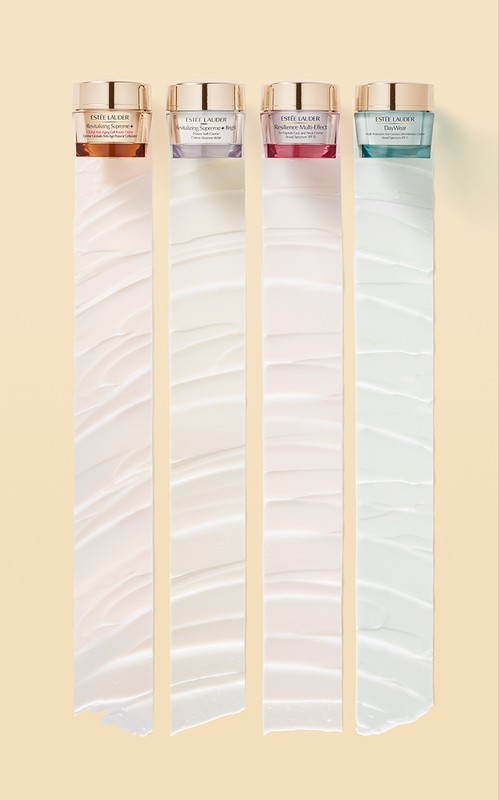 Four Estee Lauder skincare jars in shades of gold, purple, pink, and teal are arranged on a beige backdrop with ribbons of cream.