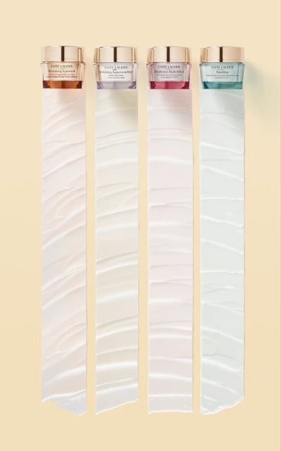 Four Estee Lauder skincare jars in shades of gold, purple, pink, and teal are arranged on a beige backdrop with ribbons of cream.