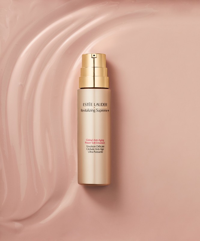 Estee Lauder Revitalizing Supreme+ emulsion bottle on a pale pink wavy background. Beauty product shot in a studio setting with soft lighting.