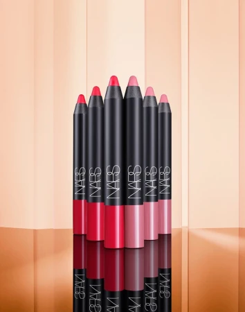 Seven NARS lip crayons stand in a row with colors ranging from bright red to light pink, positioned on a reflective surface.