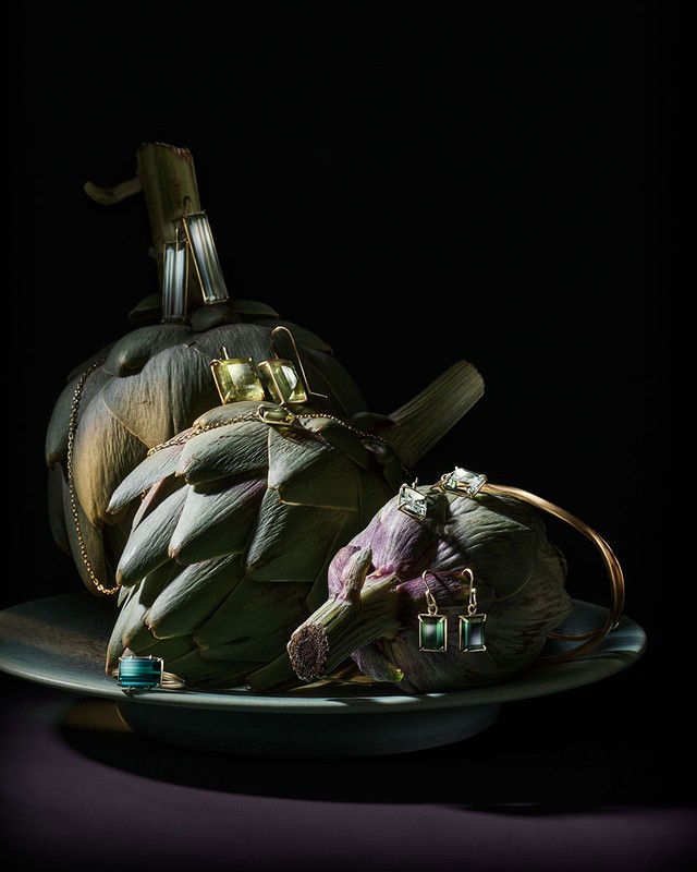 Fine jewelry displayed on artichokes, set on a gray plate against a black background. Gold, silver, and emerald tones.