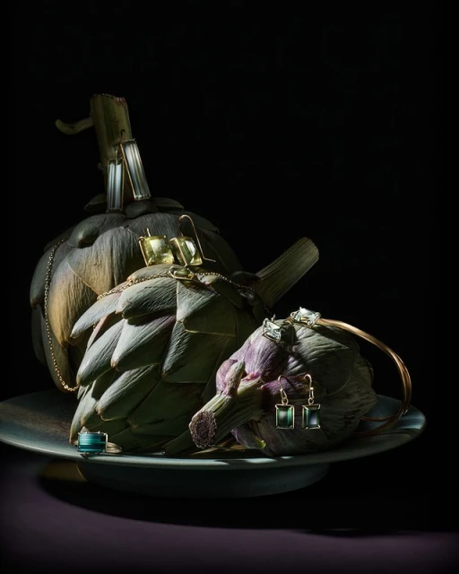Fine jewelry displayed on artichokes, set on a gray plate against a black background. Gold, silver, and emerald tones.