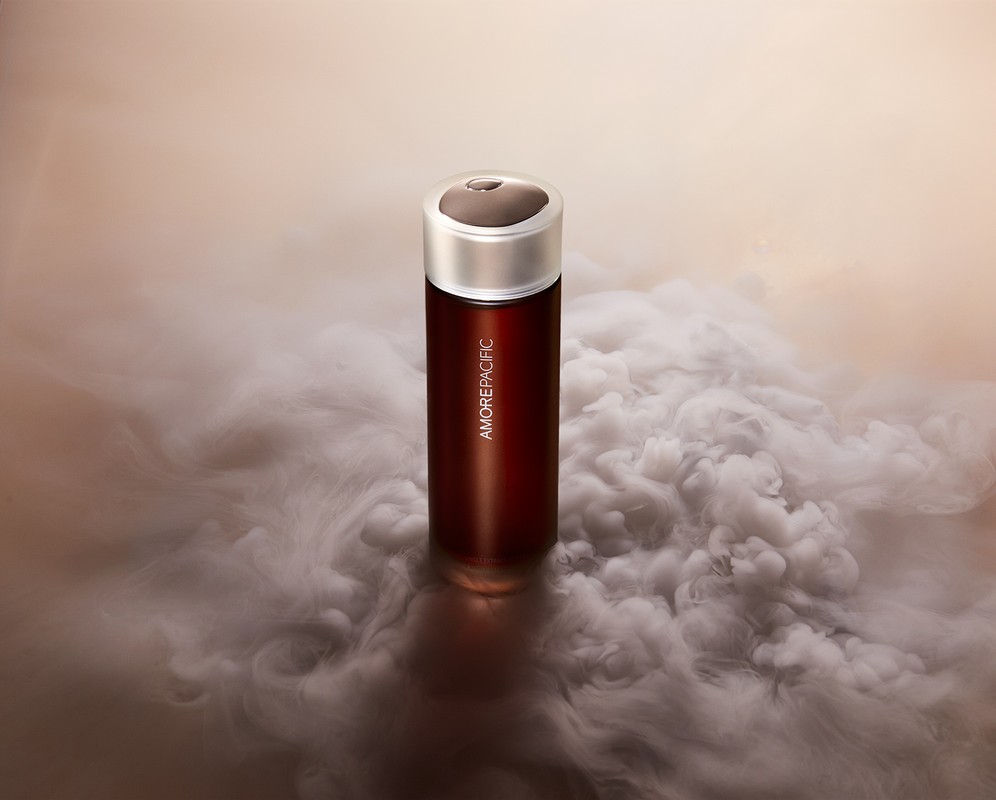 Amorepacific skincare product in a tall red bottle with a silver cap, enveloped by a cloud of smoke, on a neutral-toned surface.