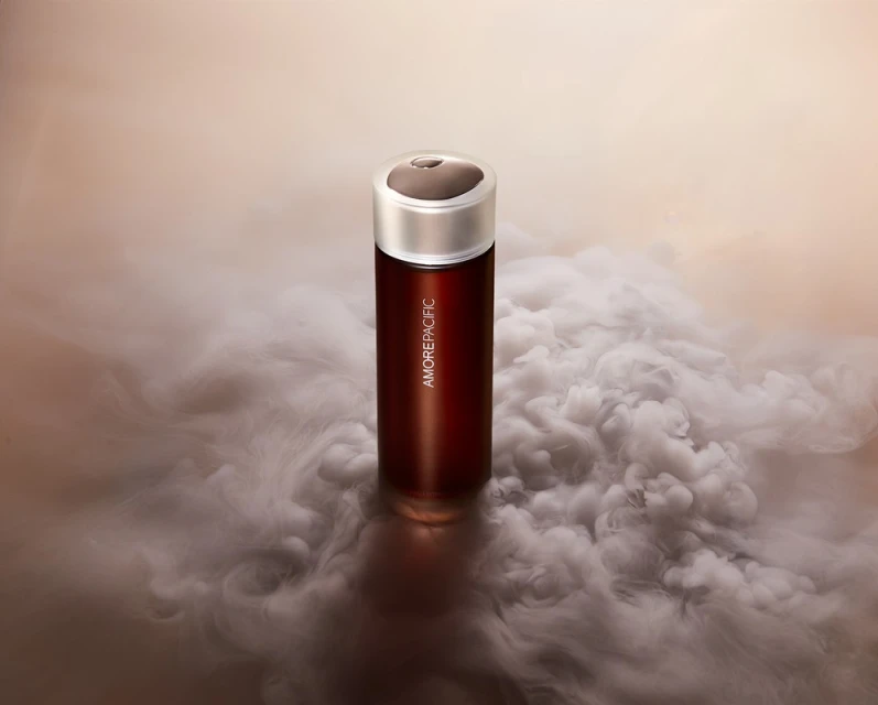 Amorepacific skincare product in a tall red bottle with a silver cap, enveloped by a cloud of smoke, on a neutral-toned surface.