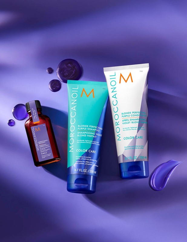 Moroccanoil Blonde Perfecting Purple Shampoo, Conditioner, and hair oil arranged against a violet backdrop with drops and smear.