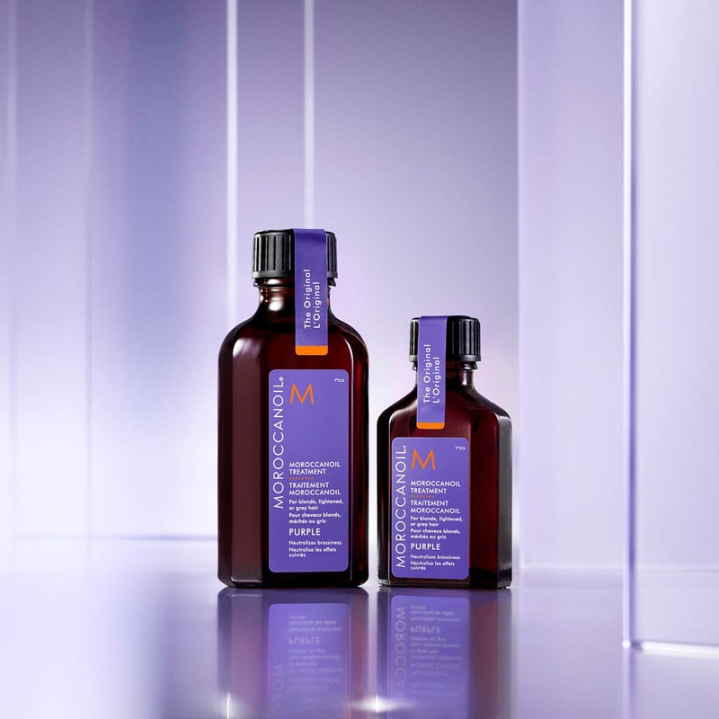 Two bottles of Moroccanoil Treatment Purple hair product on a reflective surface against a blurred purple and white background.