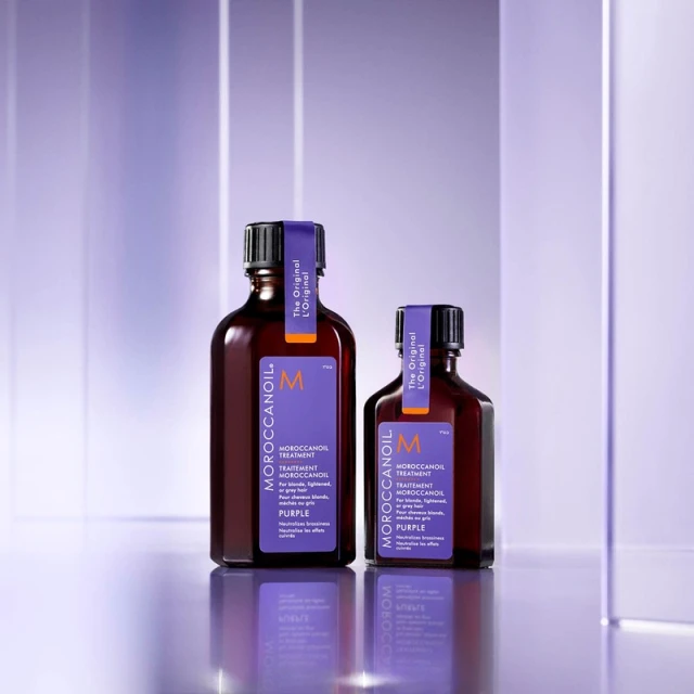 Two bottles of Moroccanoil Treatment Purple hair product on a reflective surface against a blurred purple and white background.