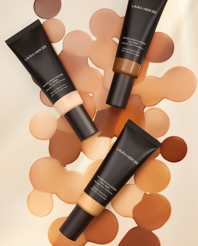 Laura Mercier tinted moisturizer tubes in various shades of tan, arranged on a light surface with spots of matching liquid.