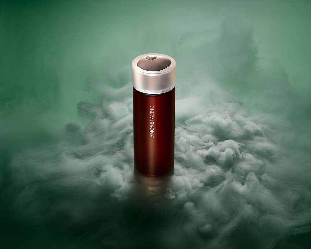 Amorepacific skincare product in a dark red container with a silver lid sits in a cloud of smoke against a green background.