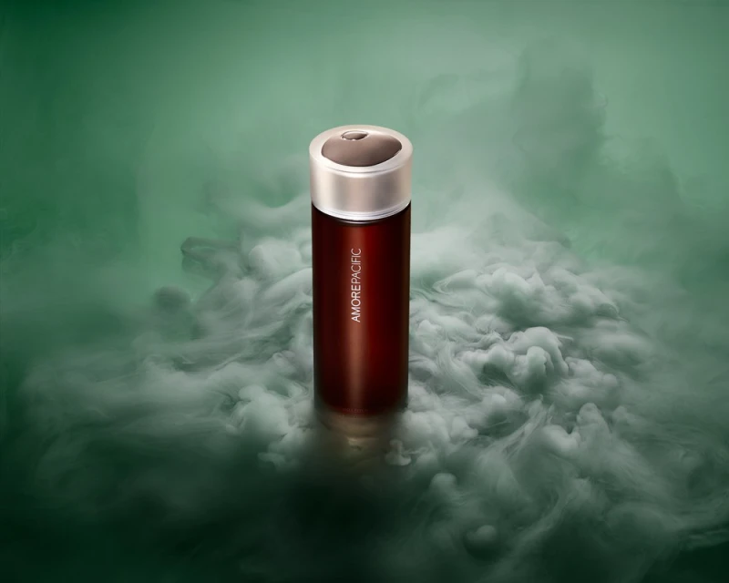 Amorepacific skincare product in a dark red container with a silver lid sits in a cloud of smoke against a green background.