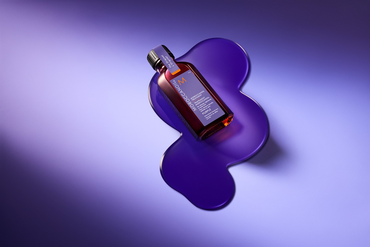 Moroccanoil purple shampoo bottle on a purple puddle against gradient blue and purple background in a beauty still life product shot.