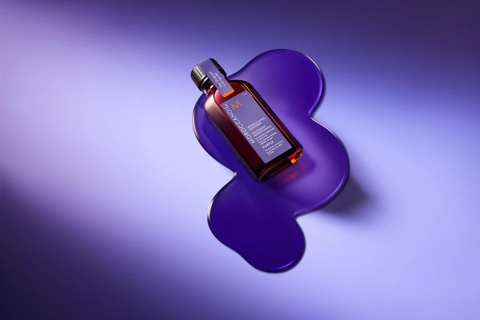 Moroccanoil purple shampoo bottle on a purple puddle against gradient blue and purple background in a beauty still life product shot.